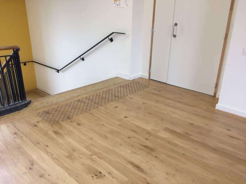 Intérieur lumineux avec parquet clair menant à des portes blanches. Zone de contraste visuel près d'un escalier avec rampe noire.