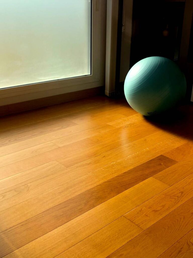 Sol en parquet clair baigné de lumière naturelle devant une fenêtre givrée. Un ballon d'exercice bleu pastel repose dans un coin.