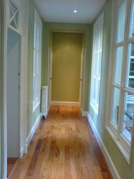 Long couloir vert clair avec parquet en bois, fenêtres blanches sur la droite et portes ouvertes au fond. Design intérieur lumineux.