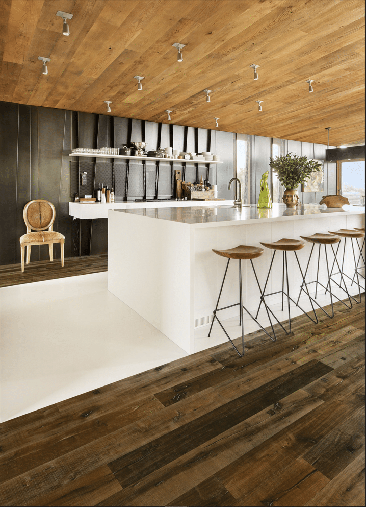 Cuisine design moderne avec grand îlot central blanc, plafond en bois et mur sombre texturé. Sol bi-matière bois foncé et résine claire. Tabourets de bar en bois et métal.