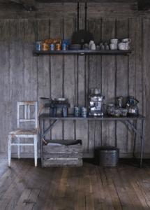 Cuisine rustique bois. Étagère: conserves, tasses. Table: réchaud, machine espresso, bouilloire. Chaise. Caisse bois texte visible.