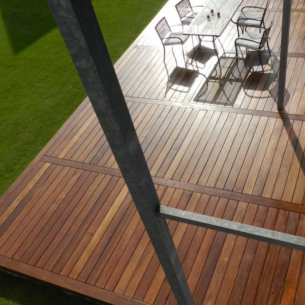 Deck en bois clair et structuré, avec table en verre et chaises de jardin sous le soleil. Poteau métallique et pelouse verte.