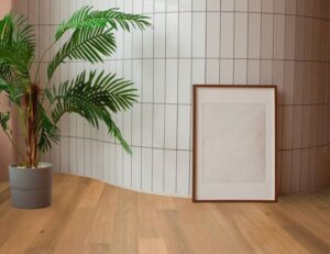 Palmier en pot devant un mur carrelé blanc incurvé, avec un cadre photo vide posé sur le parquet bois clair.