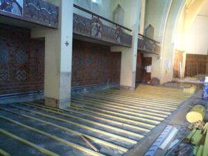 Intérieur d'église en rénovation avec sol en lattes de bois clair, piliers et murs décorés de mosaïques. Une croix rouge est sur un pilier.