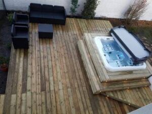 Deck en bois clair avec salon de jardin noir et table basse. Un jacuzzi moderne est encastré dans le deck, partiellement couvert.