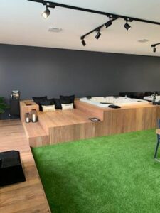 Intérieur design avec jacuzzi encastré sur plateforme en bois. Coin salon, sol gazon synthétique vert. Éclairage moderne au plafond.