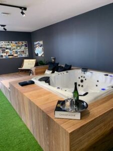 Jacuzzi blanc encastré sur estrade bois, champagne et flûtes. Espace moderne, murs gris et gazon synthétique. Boîte "CHAMPAGNE".