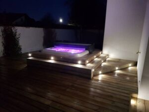 Jacuzzi illuminé en violet intégré à une terrasse en bois avec des marches éclairées, créant une ambiance chaleureuse de nuit.