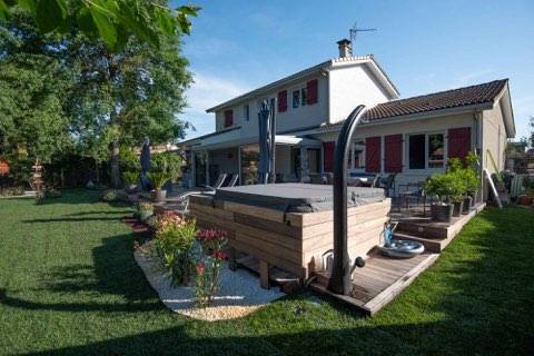 Jardin aménagé avec spa en bois intégré, douche extérieure, grande pelouse et maison moderne. Espace détente ensoleillé.