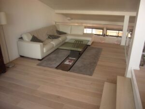 Salon loft moderne lumineux avec grand canapé d'angle blanc, table basse en verre design, tapis gris et parquet clair. Espace décloisonné.