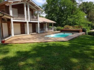 Maison en bois avec volets blancs, grande terrasse en bois et piscine rectangulaire, entourées d'un jardin verdoyant et d'arbres.