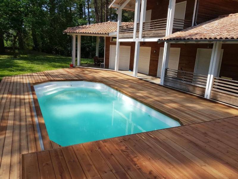 Vaste terrasse en bois clair avec piscine turquoise devant une grande maison moderne en bois à balcons et volets blancs, entourée d'arbres verdoyants.