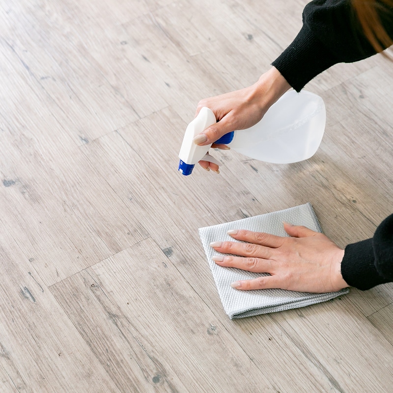 Nettoyer Efficacement Votre Plancher en Bois Une personne nettoie un plancher en bois clair avec un vaporisateur et un chiffon gris pour enlever la saleté.