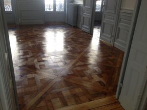 Pièce vide avec parquet bois brillant à motif géométrique complexe. Lumière du soleil sur sol ciré, portes et murs gris.