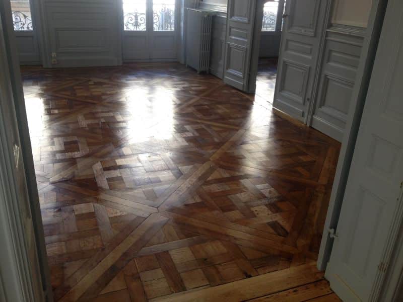 Pièce vide avec parquet bois brillant à motif géométrique complexe. Lumière du soleil sur sol ciré, portes et murs gris.
