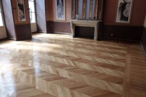 Pièce élégante avec parquet en bois clair poli motif chevrons, murs bicolores, cheminée en marbre et fenêtres lumineuses.