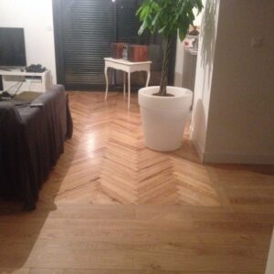 Salon lumineux avec parquet en bois clair à motif chevrons et lattes droites. Un grand pot blanc avec plante verte, un canapé et une table vintage.