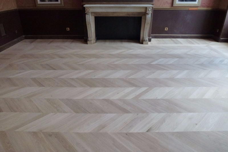 Sol en parquet chevrons de chêne clair dans une pièce élégante avec cheminée en pierre sculptée et murs sombres.