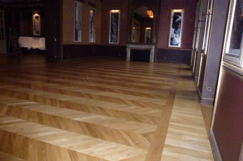 Grande salle élégante avec un parquet en bois clair à motif chevrons et bordure, murs sombres ornés de tableaux et un grand miroir doré.