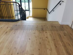 Palier en parquet clair avec bande podotactile métallique signalant un escalier. Rampes noires et main courante bois, baies vitrées.