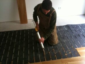 Homme posant du parquet en bois clair sur sous-plancher noir, appliquant de la colle avec un pistolet. Travaux d'installation de sol en bois.