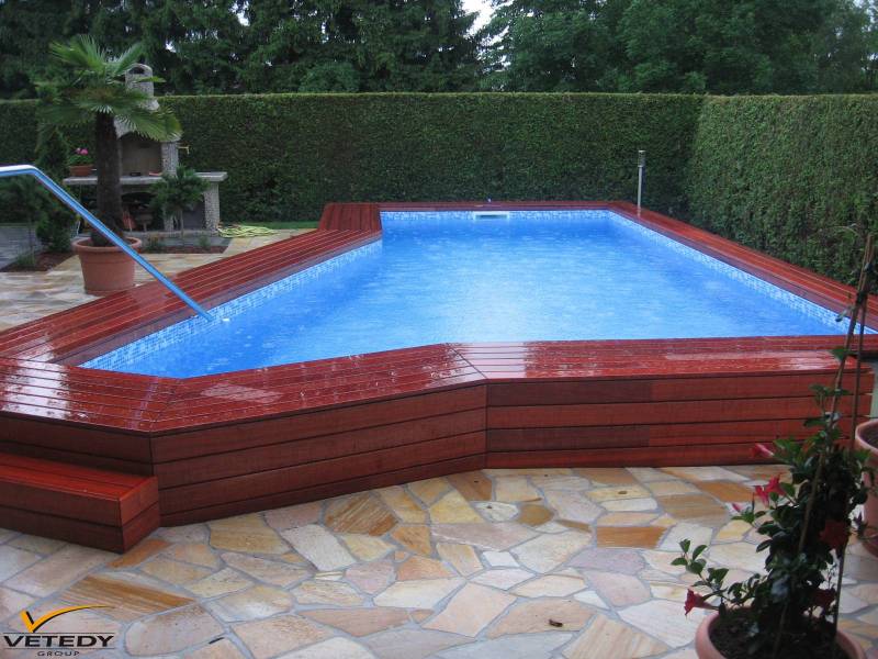 Piscine extérieure avec margelle et deck en bois rouge, carrelage bleu. Jardin aménagé avec palmier, barbecue, sol en pierre. Logo Vetedy Group.