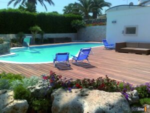 Piscine moderne avec large deck en bois et mobilier de jardin Softline. Transats bleus, canapé lounge, végétation luxuriante et palmiers.