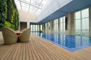 Piscine intérieure moderne avec jardin vertical luxuriant, deck en bois clair, chaises en rotin et grandes fenêtres offrant une vue sur la ville.