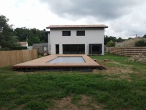 Maison moderne blanche avec piscine intégrée à une terrasse en bois, jardin en aménagement avec clôture en bambou et palissade.