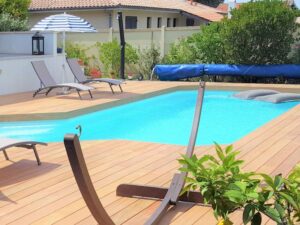 Belle piscine privée avec deck en bois clair, transats, parasol rayé et plantes. Flotteurs gris dans l'eau turquoise.