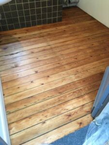 Plancher en bois clair fraîchement restauré, avec lames apparentes et veinures naturelles, près d'un mur carrelé sombre et moquette.