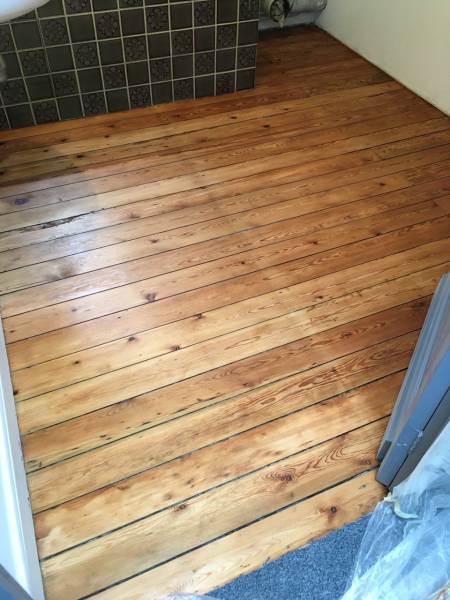 Plancher en bois clair fraîchement restauré, avec lames apparentes et veinures naturelles, près d'un mur carrelé sombre et moquette.