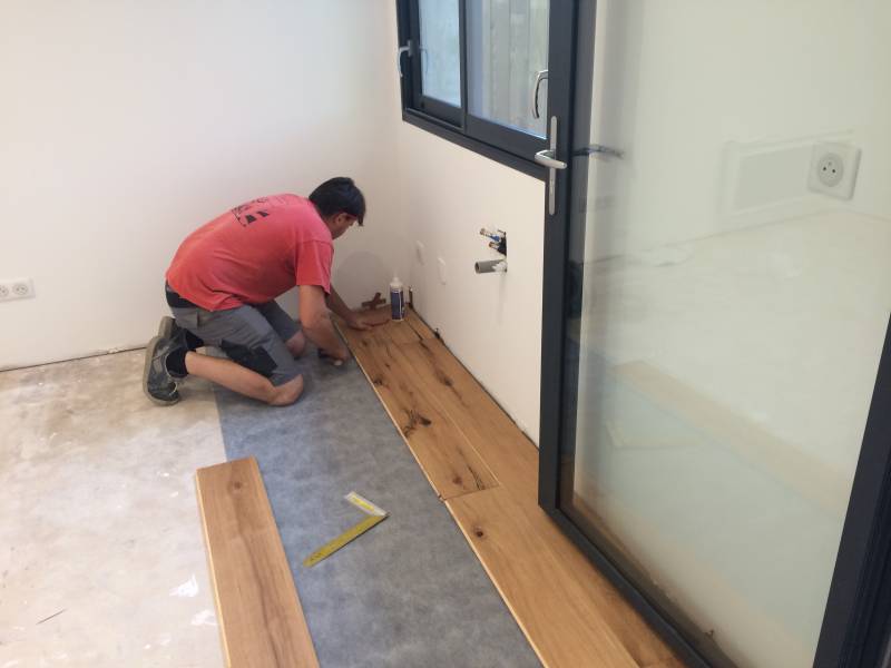 Homme agenouillé posant un parquet en bois clair sur sous-couche grise. La pièce rénovée a des murs blancs et une grande baie vitrée.