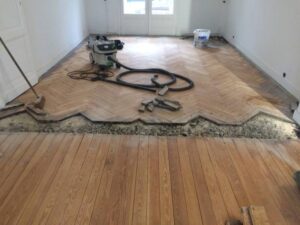 Rénovation de parquet chevrons et planches de bois. Ponceuse Festool et aspirateur au sol, une zone de parquet est retirée pour réparation.