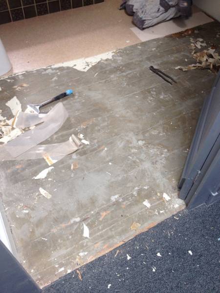 Sol de salle de bain en rénovation: revêtement ancien retiré, laissant apparaître sous-plancher gris et débris. Outils de démolition visibles.