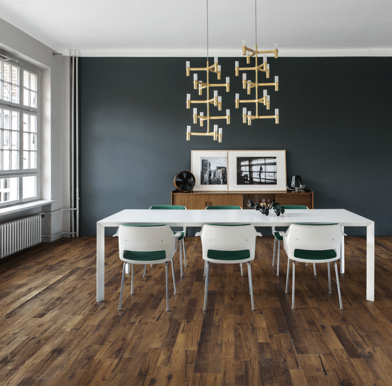 Salle à manger moderne avec grande table blanche, chaises design vertes, parquet rustique, buffet en bois et lustre doré sculptural.