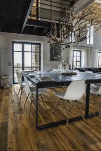 Salle à manger industrielle loft, grande table en bois, chaises modernes, lustre branches, parquet chêne, lumière naturelle.