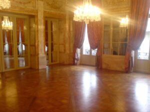 Somptueux salon de bal vide avec parquet brillant, hauts plafonds dorés, et grands lustres en cristal. Fenêtres drapées de rideaux rouges.