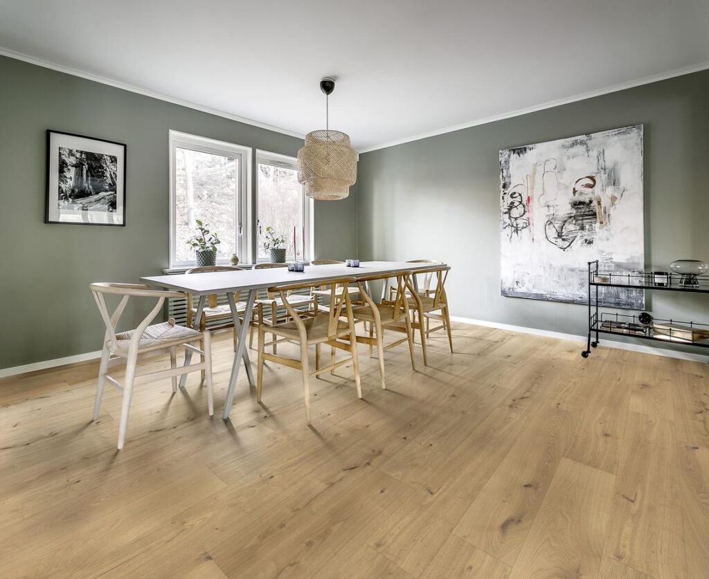 Salle à manger scandinave moderne avec murs vert sauge, parquet bois clair, table blanche et chaises Wishbone, éclairée par un lustre en rotin.