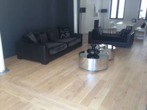 Salon moderne: canapés noirs, coussin 'LOVE', tables basses rondes argentées et parquet clair à chevrons. Espace repas en fond.