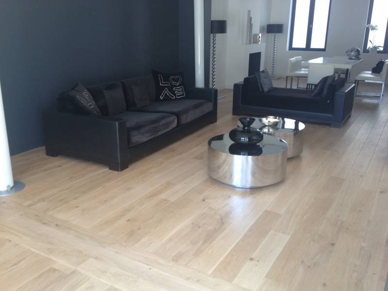 Salon moderne: canapés noirs, coussin 'LOVE', tables basses rondes argentées et parquet clair à chevrons. Espace repas en fond.