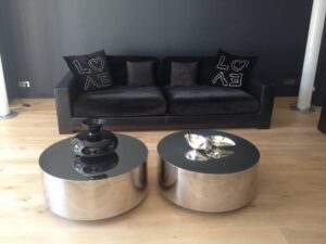 Salon moderne: canapé noir avec coussins "LOVE", deux tables basses rondes argentées et décoration chic sur parquet clair.