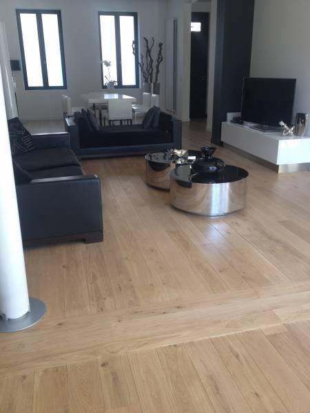 Salon moderne avec canapé noir, tables basses rondes chromées, TV, et parquet clair. Grandes fenêtres et salle à manger en arrière-plan.