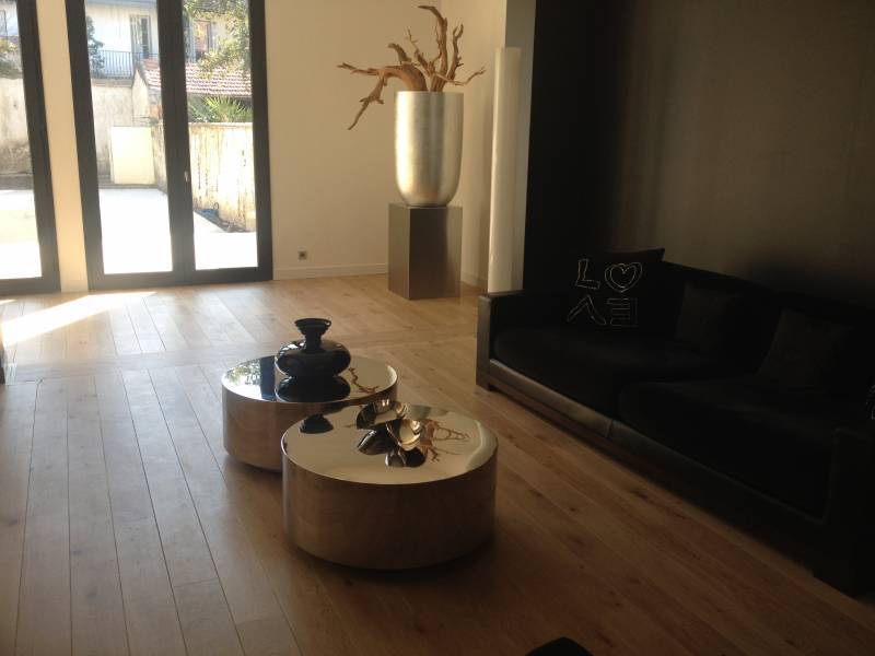 Salon moderne lumineux avec parquet clair. Deux tables basses rondes, un canapé noir (coussin "LOVE"), grand vase et branches décoratives.