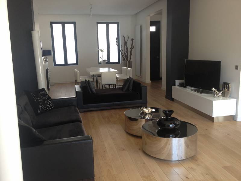 Salon moderne noir et blanc avec canapés foncés et tables basses chromées. Salle à manger lumineuse en fond, fenêtres noires. Coussin 'LOVE'.