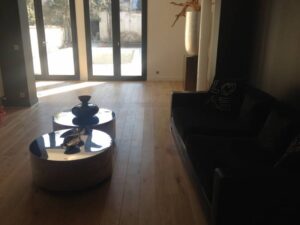 Salon lumineux avec parquet clair, deux tables basses rondes réfléchissantes, canapé sombre marqué 'LOVE' et grandes portes vitrées donnant sur un patio.