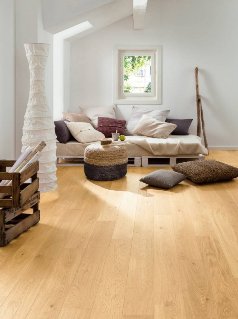 Salon lumineux avec parquet clair et canapé palettes. Coussins, pouf tressé, lampe papier et caisse "Wein aus deutschen Landen" en bois.