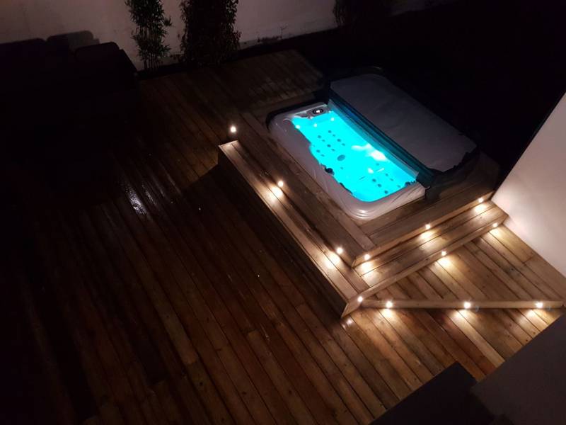 Spa lumineux bleu intégré à une terrasse en bois clair, avec des marches éclairées par des LED, la nuit. Ambiance relaxante.