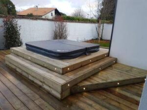 Spa extérieur avec couverture noire, installé sur une terrasse en bois à plusieurs niveaux, éclairée par des spots LED intégrés aux marches.