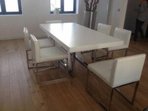 Salle à manger moderne avec grande table blanche et huit chaises rembourrées claires, sur parquet. Fenêtres lumineuses.